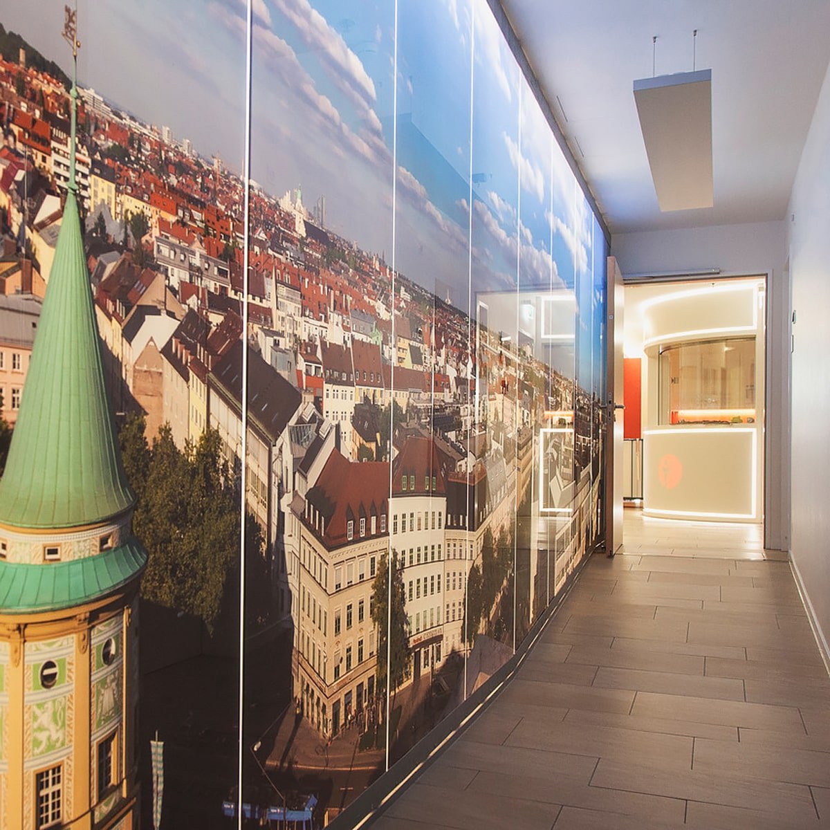 Ein Flur mit gefliestem Boden im Ortho Health Institut in München ist mit einem großen Wandgemälde geschmückt, das eine Luftaufnahme einer Stadtlandschaft mit einem Gebäude mit grüner Kuppel im Vordergrund zeigt. Am Ende des Flurs befindet sich ein gut beleuchteter Raum mit modernem Dekor und weißen und roten Akzenten.