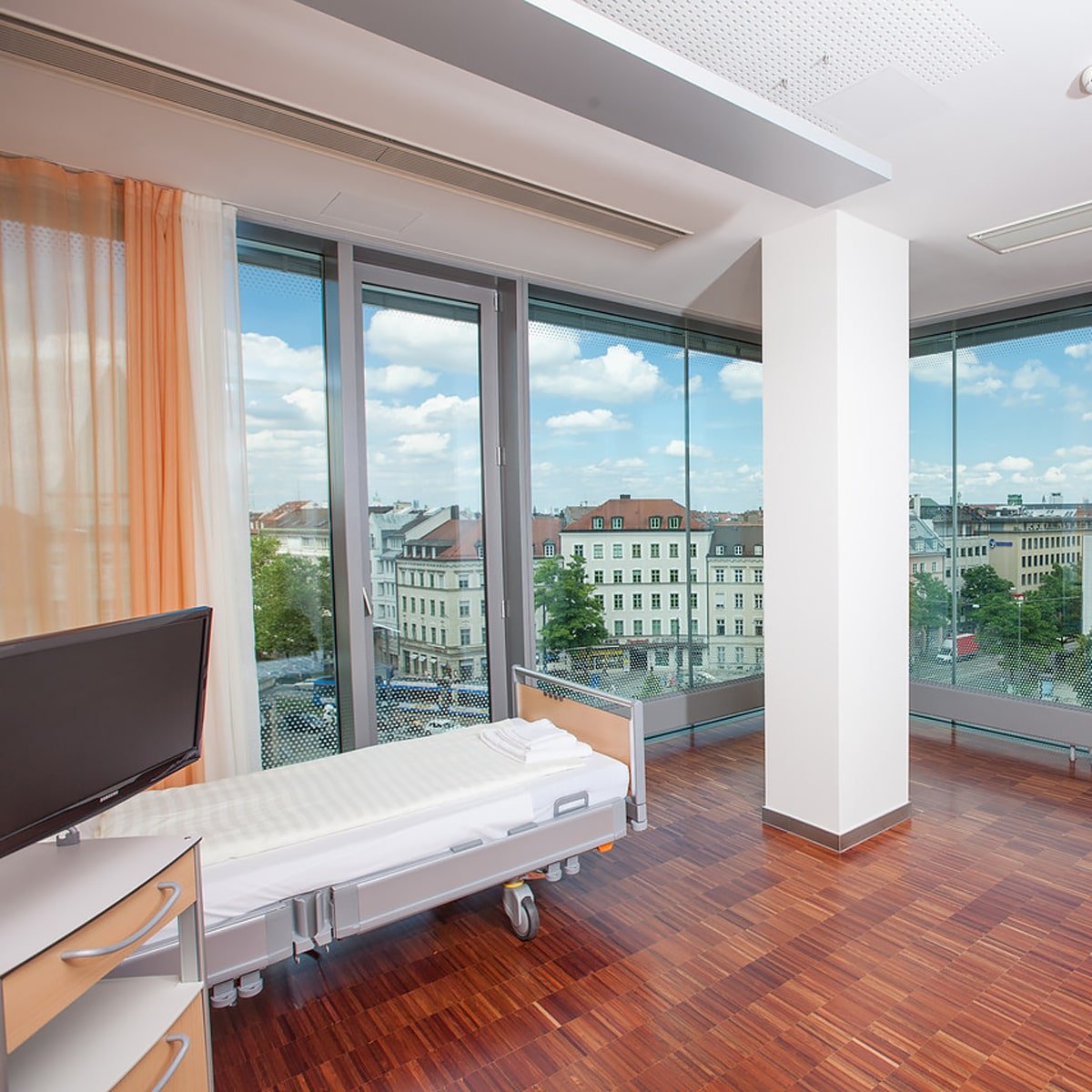 Ein modernes Krankenhauszimmer im Ortho Health Institut in München verfügt über ein Einzelbett, einen Flachbildfernseher und große, vom Boden bis zur Decke reichende Fenster mit Blick auf die Stadt mit Gebäuden und einem blauen Himmel mit Wolken. Das Zimmer hat Holzfußboden und teilweise zugezogene helle Vorhänge.