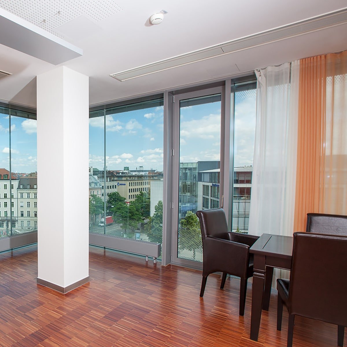 Iatros Klinik - Ein moderner Büroraum in München mit großen, deckenhohen Fenstern mit Blick auf die Stadt. Der Raum verfügt über einen kleinen Sitzbereich mit zwei dunkelbraunen Lederstühlen und einem passenden Tisch auf einem Hartholzboden. Durchsichtige Vorhänge verdecken die Fenster teilweise und sorgen für eine einladende Atmosphäre im Ortho Health Institut.